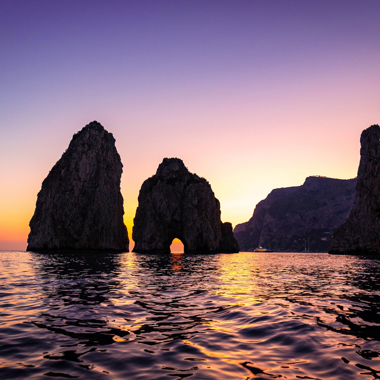 The famous Capri Faraglioni rock formations in the water during a sunset with a purple and orange sky.