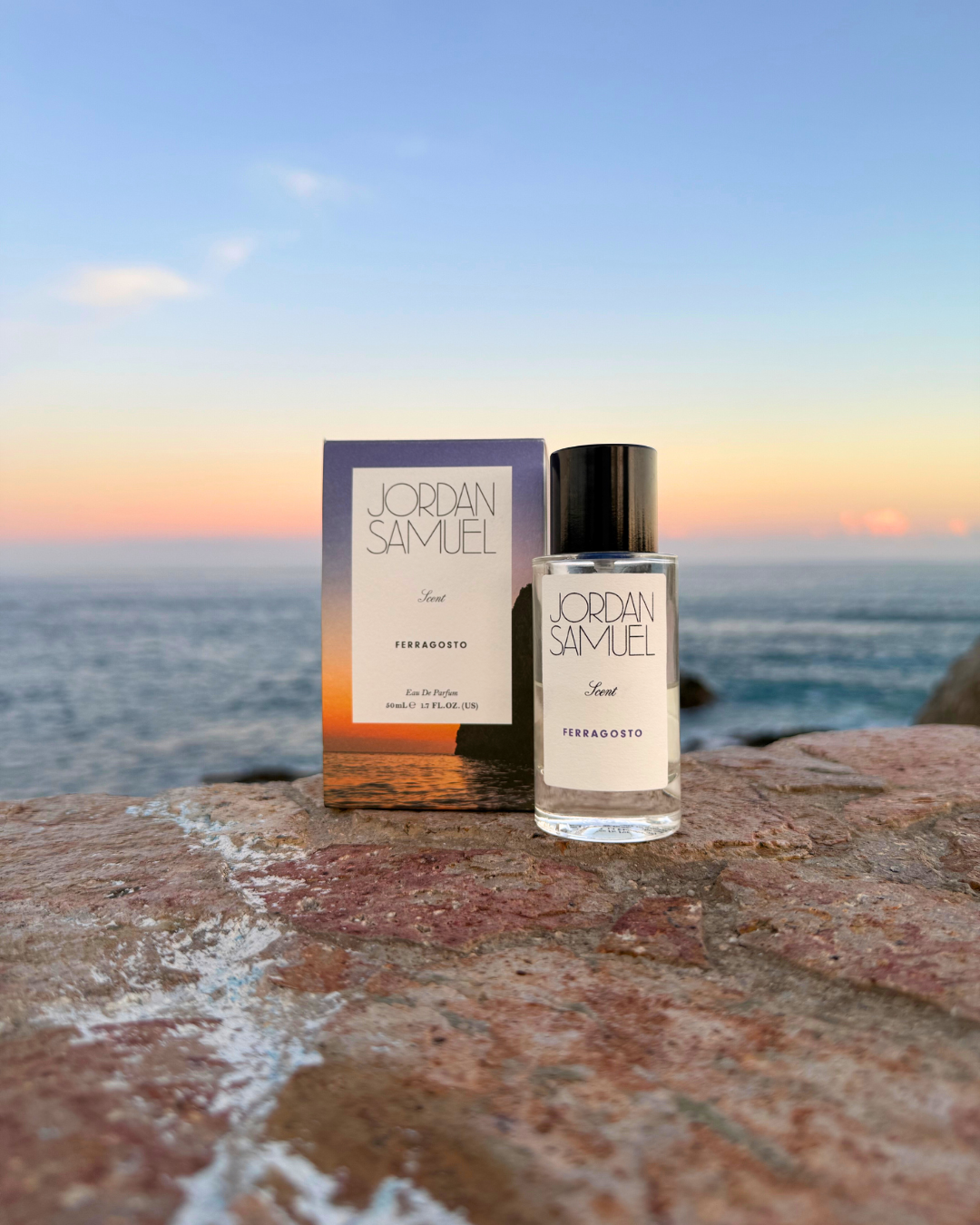 Jordan Samuel Ferrogosto perfume bottle and box packaging on a rocky surface with a scenic coastal background as the sun begins to set