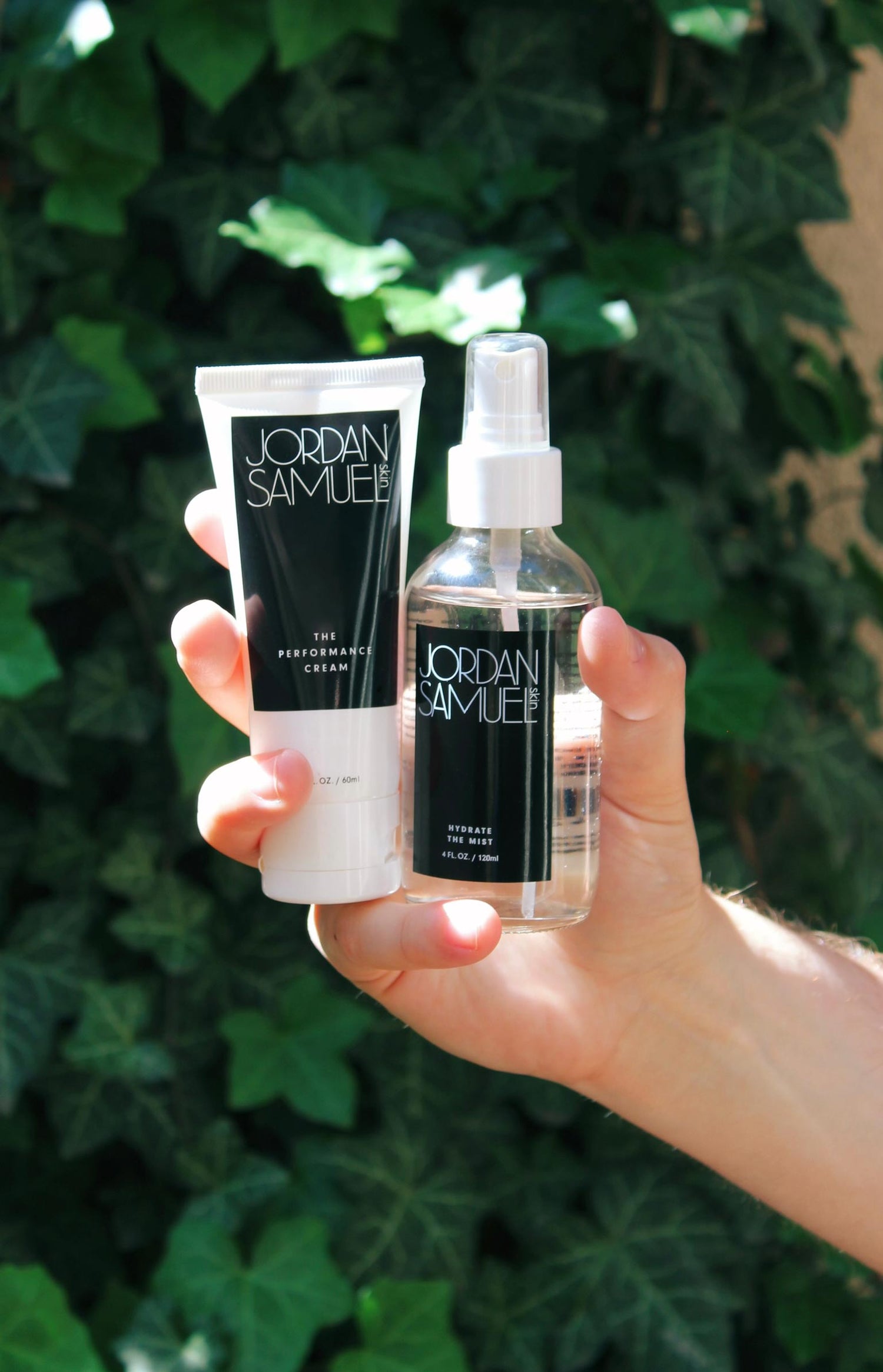Hand holding The Performance Cream's white tube with black label and Hydrate the Mist's clear glass bottle with black label up against a wall covered in green ivy.