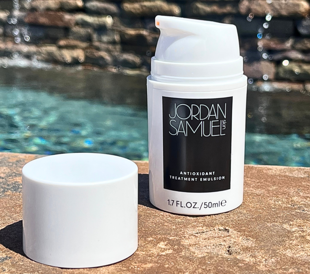Antioxidant Treatment Emulsion's white pump bottle with cap removed and sitting next to it on the ledge of a water feature. Turquoise water and a stone wall with cascading water can be seen in the background.