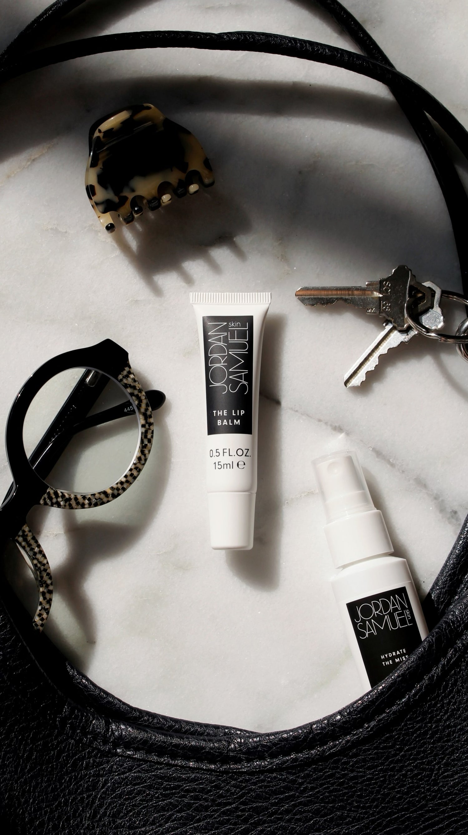 The Lip Balm's white tube with black label centered among black leather purse straps, silver keys, black and mother of pearl eyeglasses and a tortoise shell hair lip.