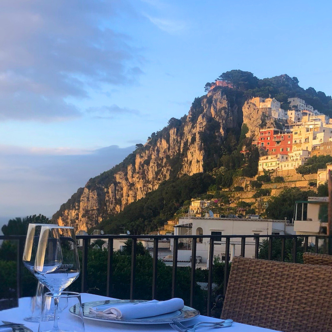 Dining table at Panorama Ristorante in Capri set with glasses and napkins overlooking a mountainous landscape with buildings.