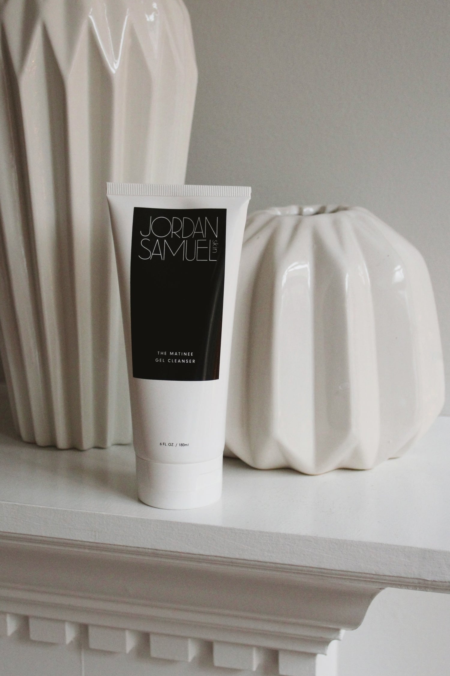 The Matinee Gel Cleanser's white tube with black label stands on a white fireplace mantle among white ceramic pottery. 