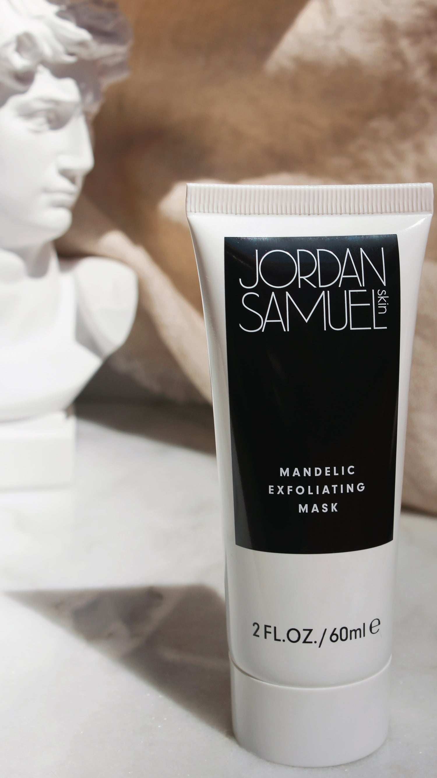 The Mandelic Exfoliating Mask's white tube with black label stands n the foreground. A white sculptural bust of a male head is blurred in the background.