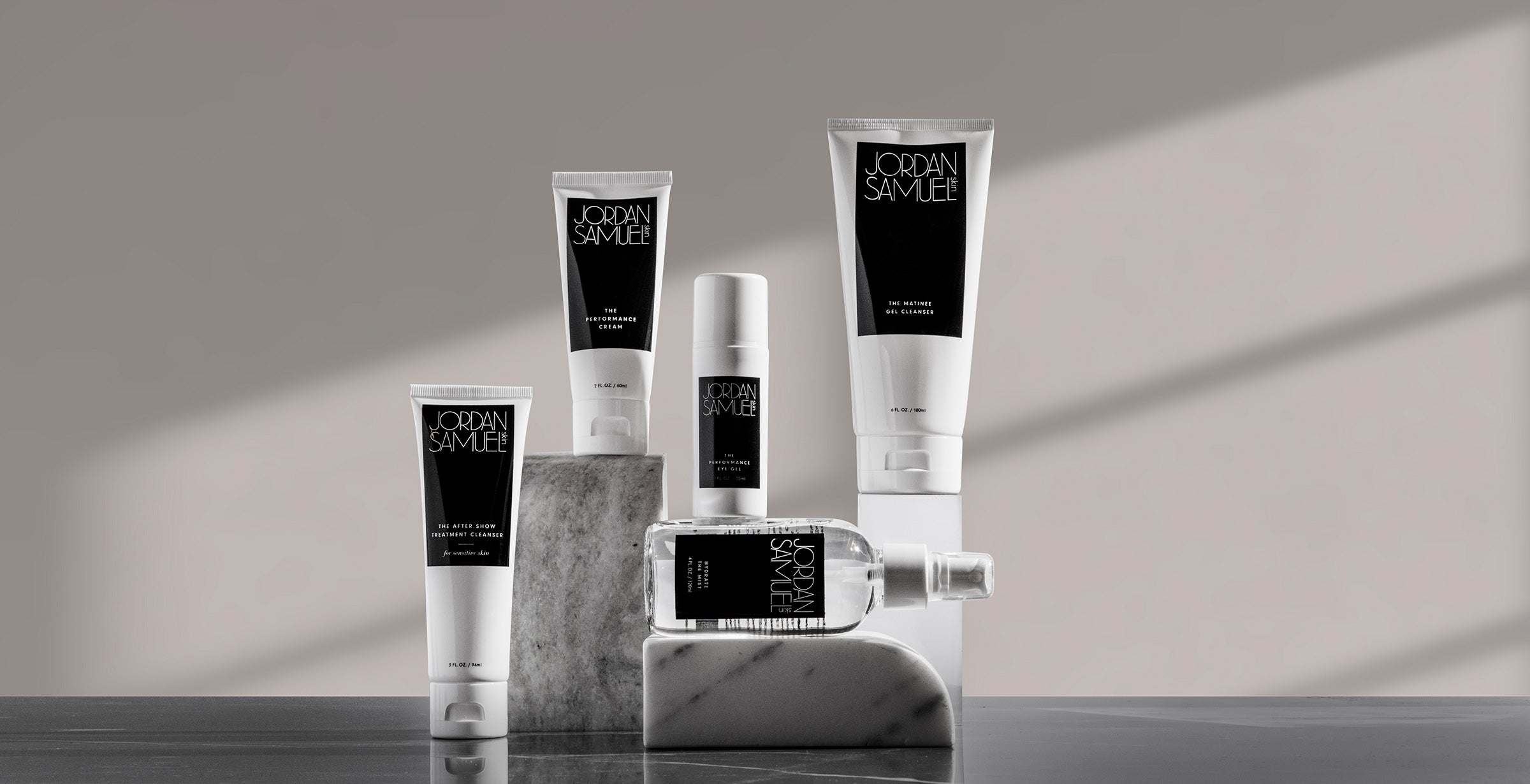 5 Jordan Samuel Skin products are arranged on white marble blocks set on a black surface. Light streams in from the window creating diagonally placed rectangle of light on the wall behind the products. 