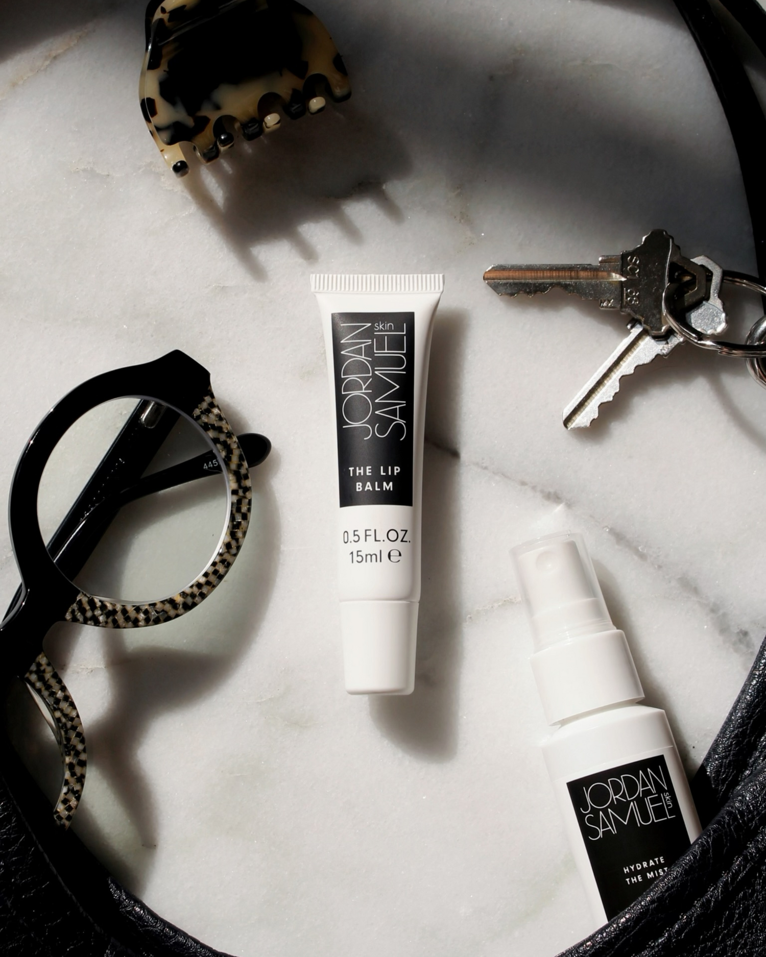 The Lip Balm's white tube with black label centered among black leather purse straps, silver keys, black and mother of pearl eyeglasses and a tortoise shell hair lip. 