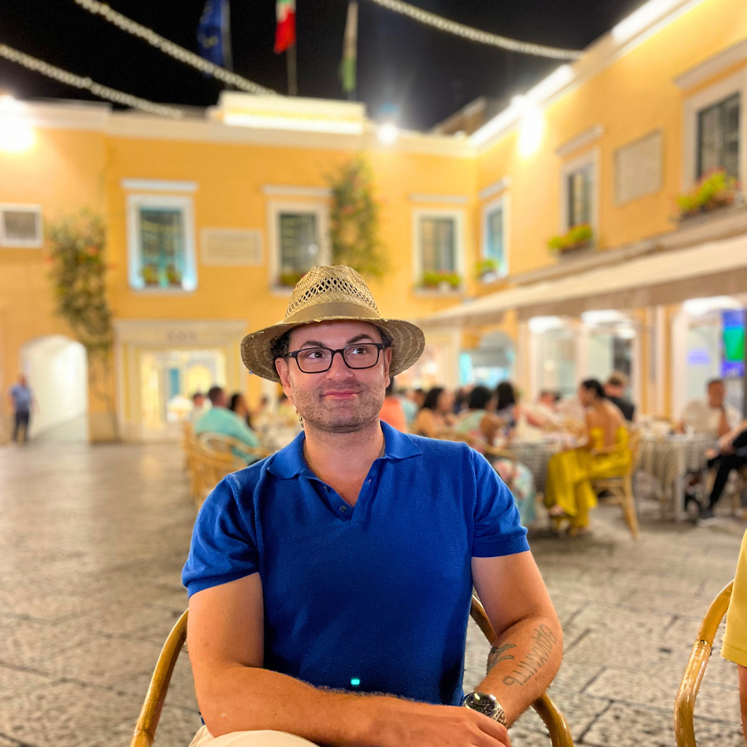 Jordan wearing a straw hat and blue shirt sitting in an outdoor Caprese cafe with a yellow building in the background