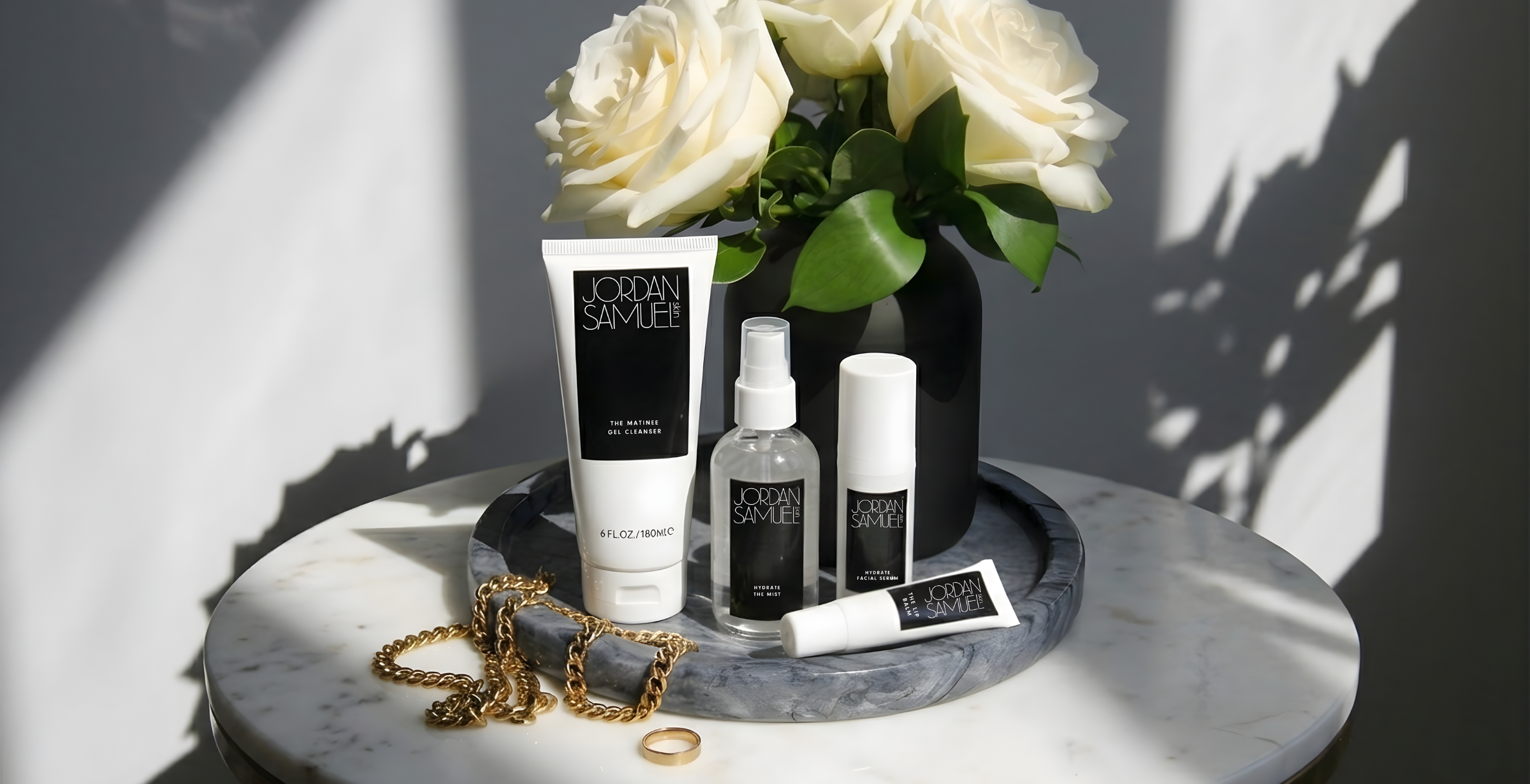 Image of The Matinee Gel Cleanser, Hydrate the Mist, Hydrate Facial Serum and The Lip Balm arranged on a gray marble tray with gold jewelry and a frosted green vase of white roses. 