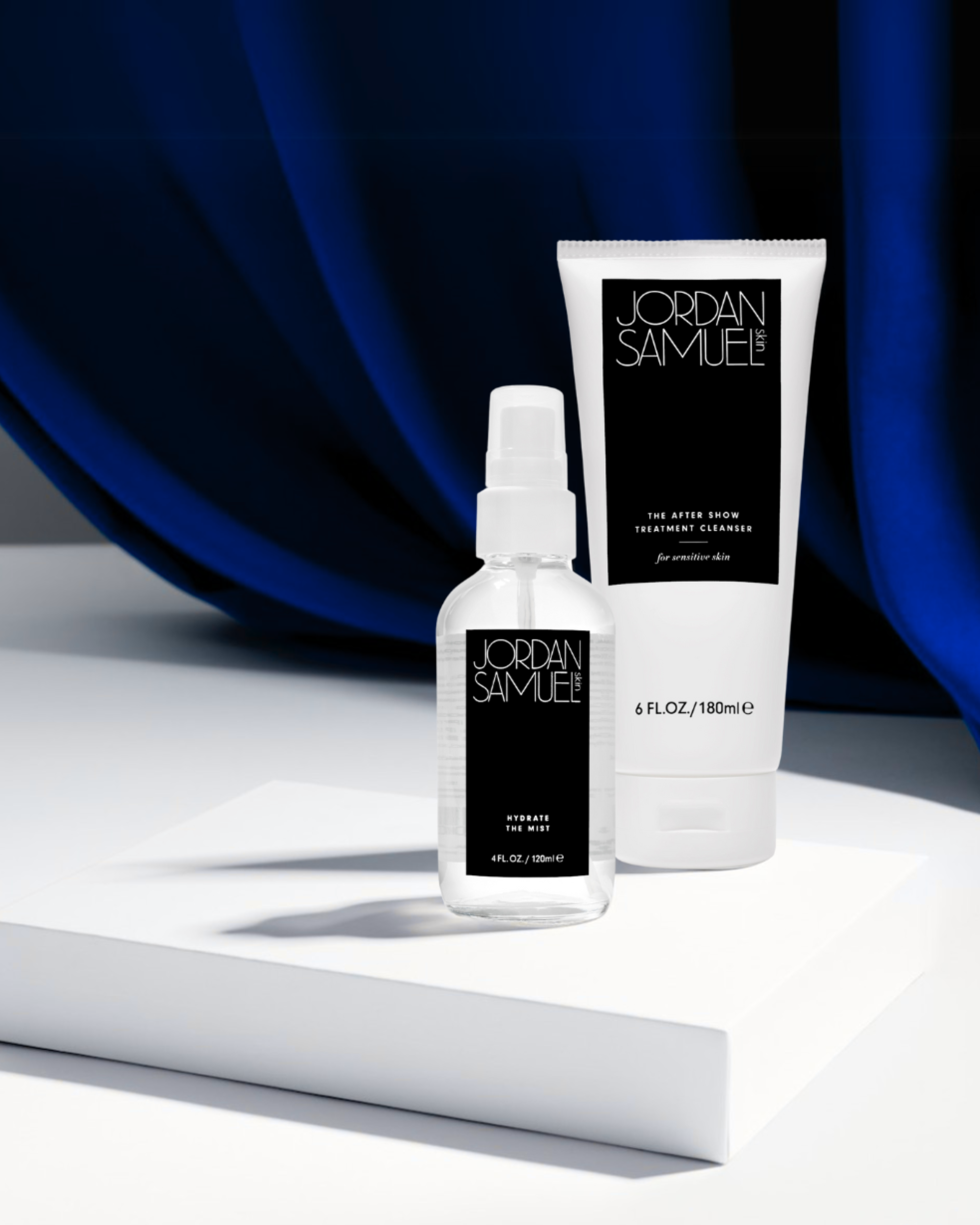 Image of The After Show Treatment Cleaner for Sensitive Skin's white tube with black label and Hydrate the Mist's clear glass bottle with black label standing on a white platform against a background of a sweeping vibrant blue velvet curtain.